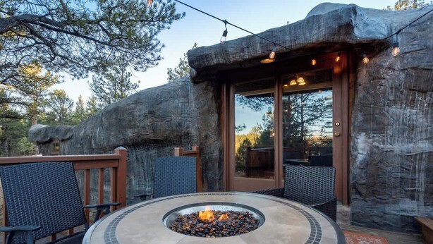A circular fire pit with a mosaic border, surrounded by wicker chairs and a wooden deck. - Cave Cabin | Hot Tub, Fire Pit & Lake (Florissant)