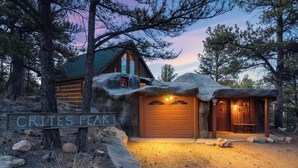 A rustic cabin with a large wooden door and a covered porch, surrounded by trees and a sign reading "CRITES PEAK". - Cave Cabin | Hot Tub, Fire Pit & Lake (Florissant)