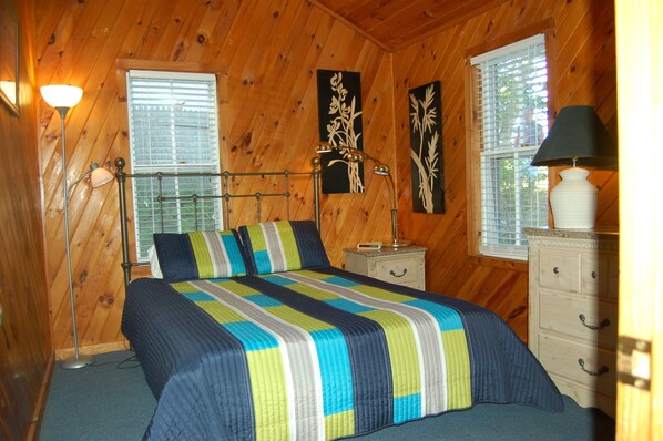 A wooden-paneled bedroom with a bed, bedside lamps, a dresser, and artwork on the walls.