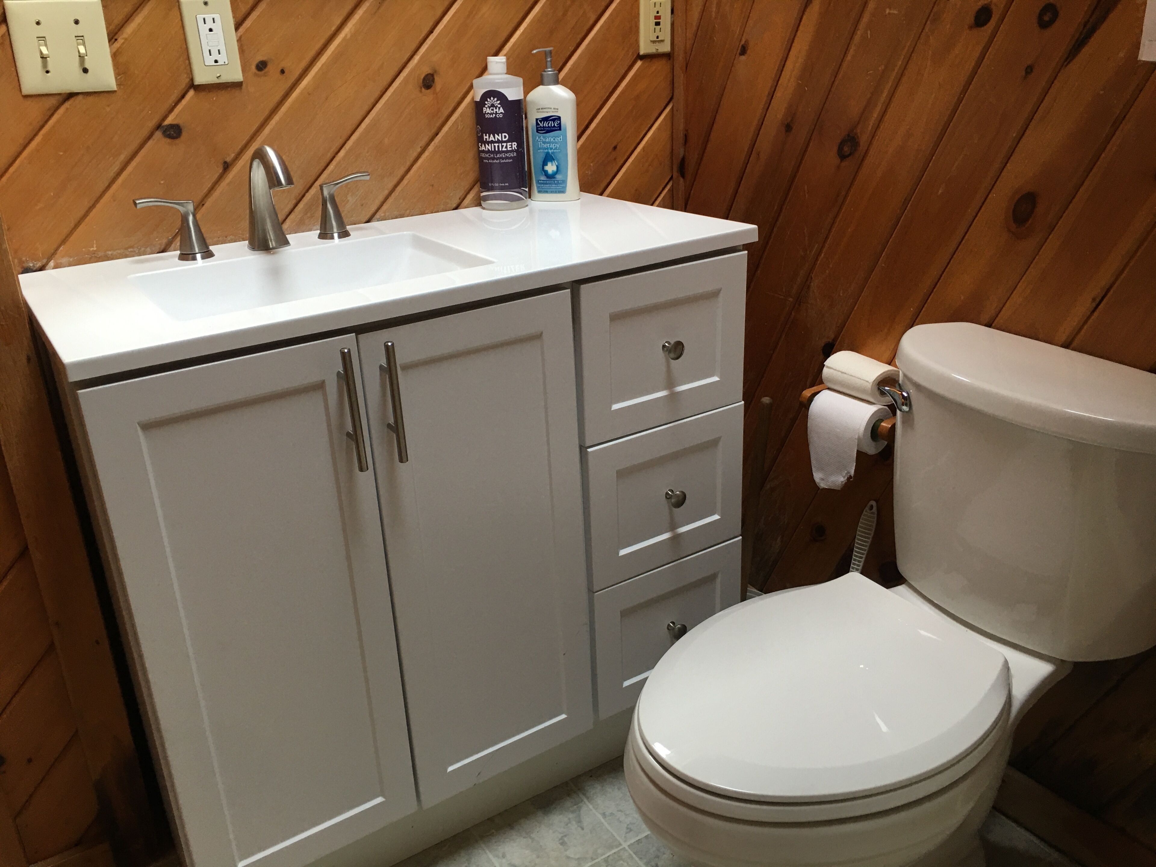 Un baño con lavamanos blanco, dos grifos, un inodoro y un mueble con cajones.