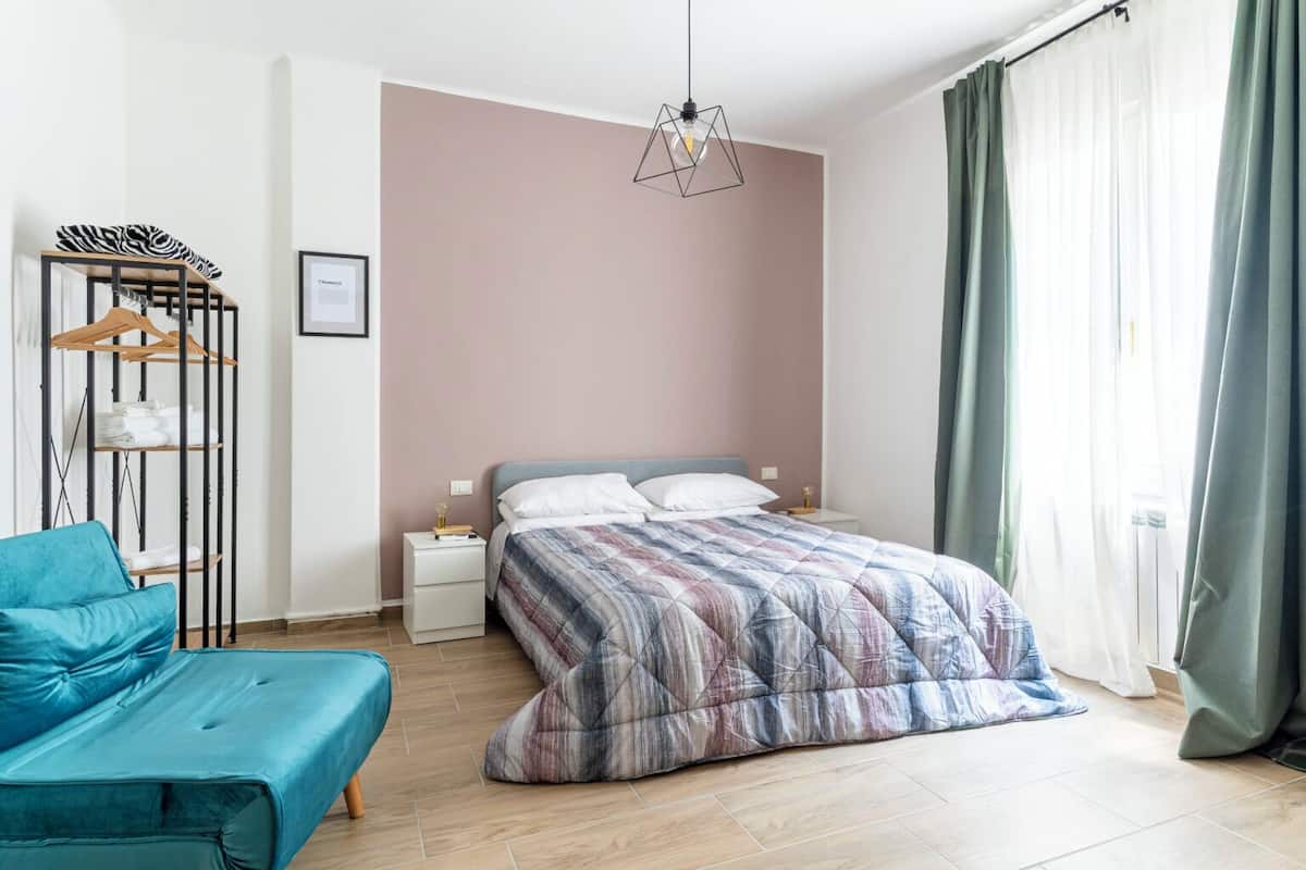 Un dormitorio con cama, mesita de noche, una silla azul, armario y una lámpara colgante.