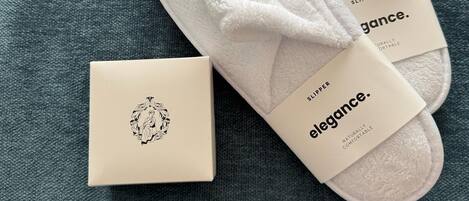 Un par de pantuflas blancas con una etiqueta que dice "elegance" y una caja pequeña con un diseño decorativo.