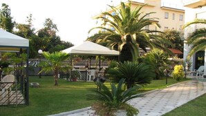 A courtyard with a paved walkway, outdoor seating, and palm trees. - Hotel Harmony&Smile (Lido di Camaiore)