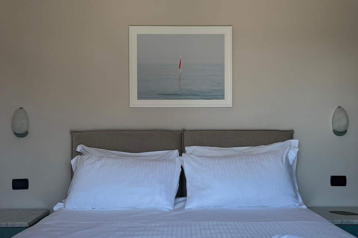 Una cama bien hecha con sábanas y almohadas blancas, un cuadro enmarcado de un velero en la pared y dos lámparas de noche.