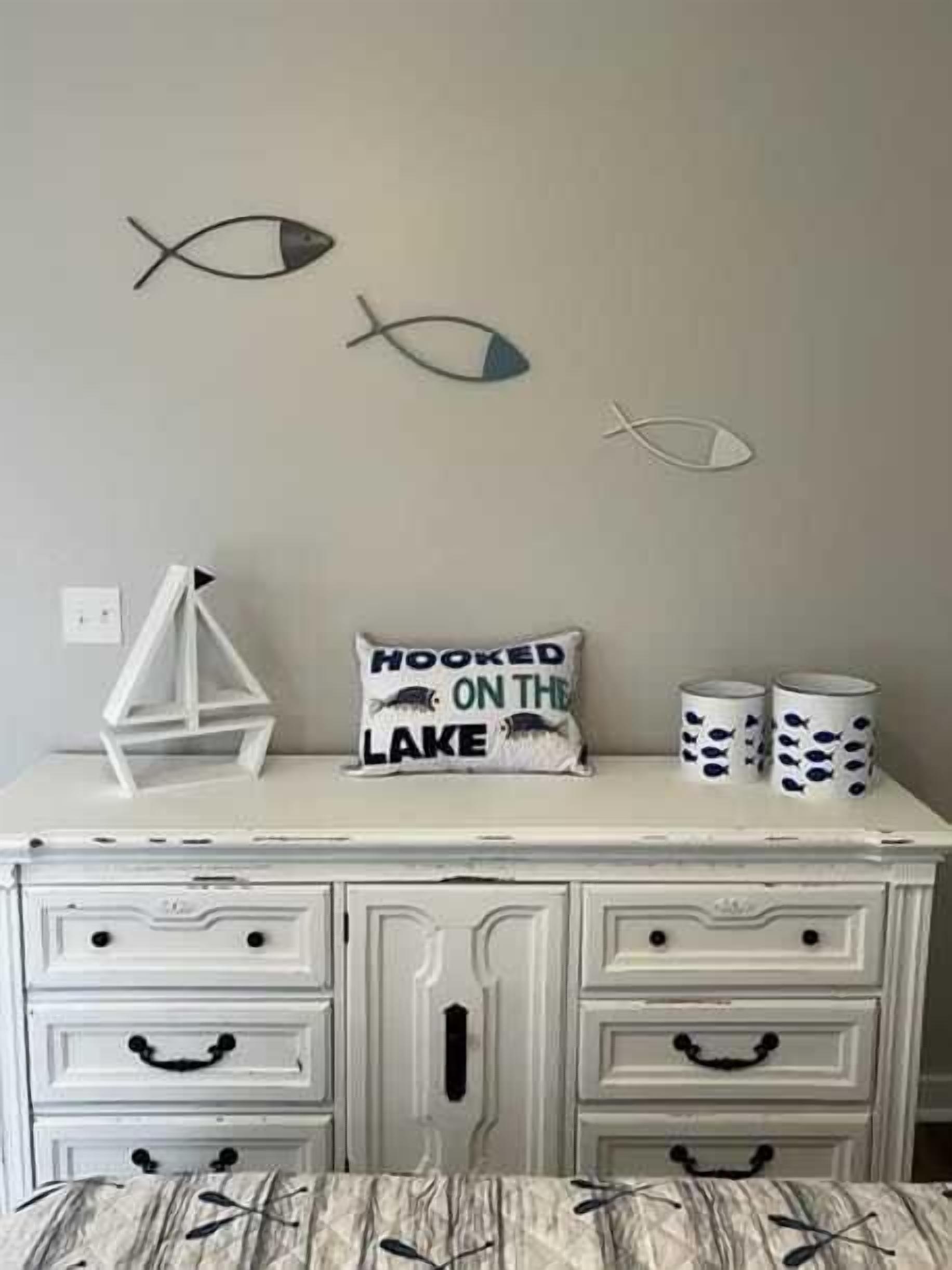 Une pièce avec une commode blanche, deux tableaux représentant des poissons, un coussin orné de l’inscription « HOOKED ON THE LAKE » et deux tasses décorées de motifs de poissons.