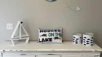 Une pièce avec une commode blanche, deux tableaux représentant des poissons, un coussin orné de l’inscription « HOOKED ON THE LAKE » et deux tasses décorées de motifs de poissons.