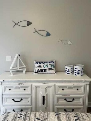A room with a white dresser, two fish wall art, a pillow with 'HOOKED ON THE LAKE' text, and two mugs with fish designs.