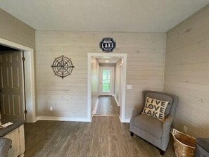 A hallway with a wooden floor, a sign reading "Welcome to Lake House," a compass wall decoration, and a grey armchair with a cushion that says "Live Love Lake."