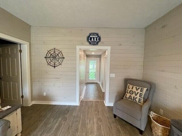 Un couloir doté d’un plancher en bois, une pancarte indiquant « Bienvenue à Lake House », une décoration murale en forme de boussole et un fauteuil gris agrémenté d’un coussin sur lequel est brodé le message « Vivez, aimez, profitez du lac ».