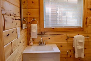 Ein Badezimmer einer Holzhütte mit einem weißen Waschbecken, einem Spiegel und einem Fenster mit Jalousien.