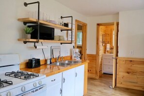 Eine kompakte Küche mit Gasherd, Mikrowelle und Kaffeemaschine. Über der Spüle befinden sich Holzregale und eine Holztür führt in einen weiteren Raum.