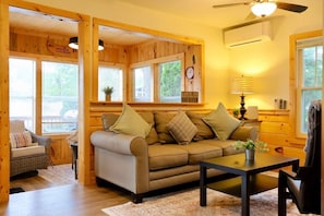 Interior acogedor de una cabaña con sofá, mesa y silla. Se observan ventanas con persianas, un ventilador de techo y un reloj en la pared.