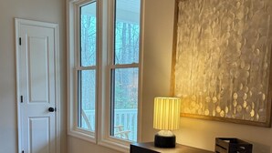 A room with a wooden floor, a white door, a window with a view of trees, a dark wooden console table with a lamp, and a textured artwork on the wall. - Charming 3-bedroom house in Louisa within 2 miles of town. (Louisa)