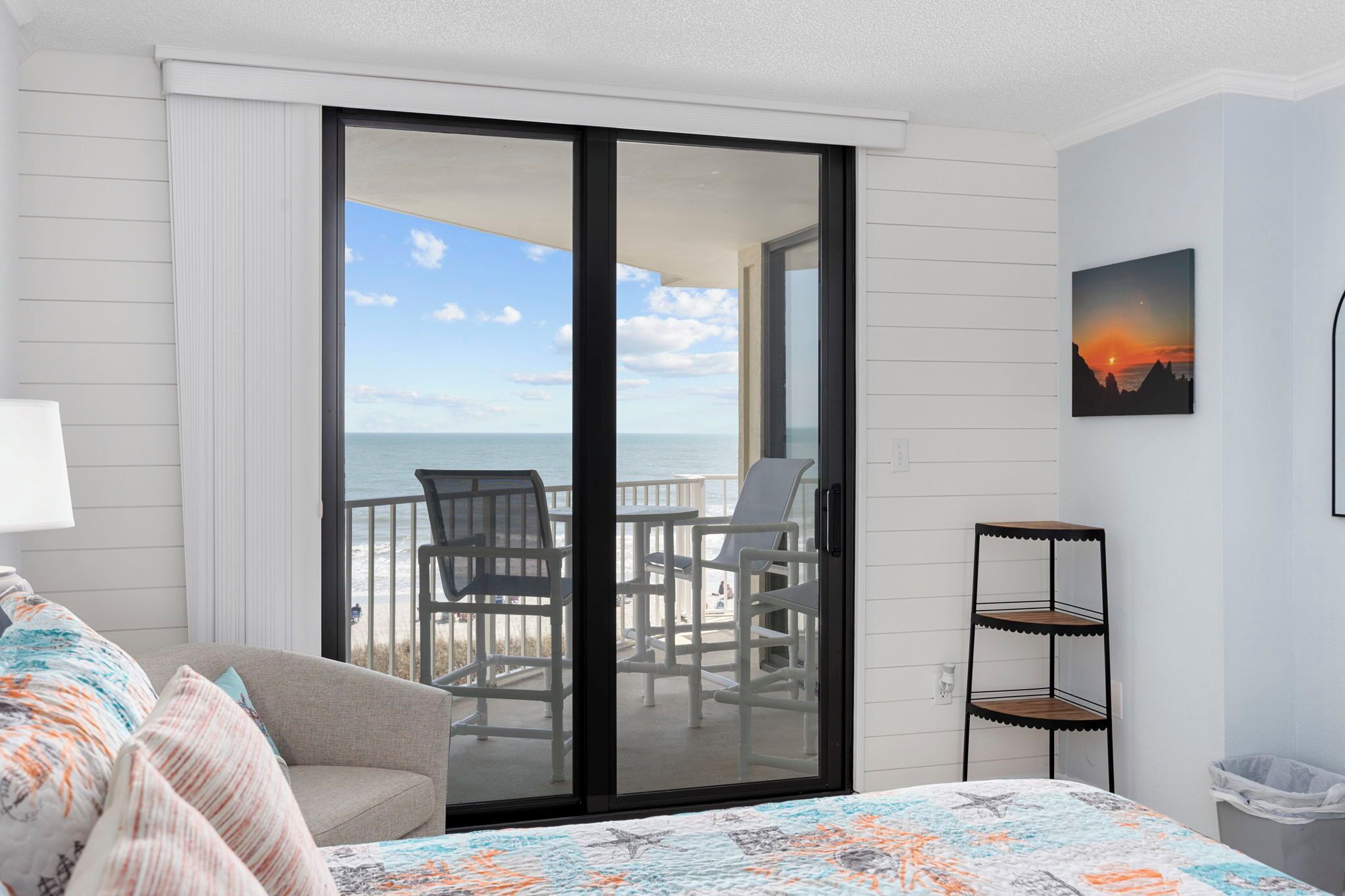 Une chambre à coucher avec un lit, une chaise, une petite table, un balcon avec des chaises et un tableau.