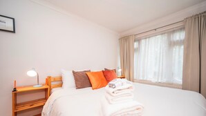 A neatly made bed with white linens and a stack of folded towels, a wooden nightstand with a lamp, and a window with sheer curtains.