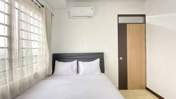 A neatly made bed with white linens in a room with a window, a wall-mounted air conditioner, and a door.
