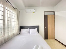 A neatly made bed with white linens in a room with a window, a wall-mounted air conditioner, and a door.