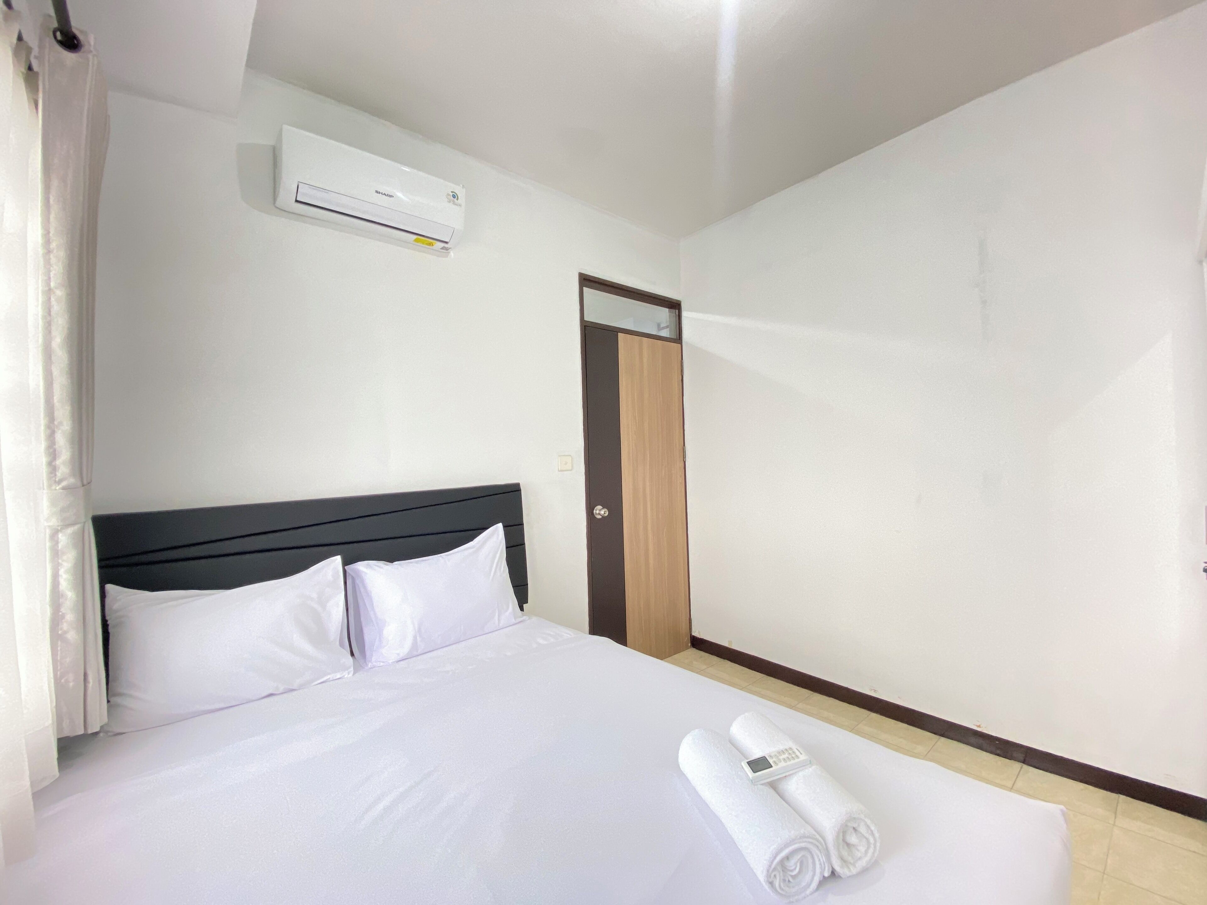 A hotel room with a bed, white linens, a digital control panel, and an air conditioning unit.