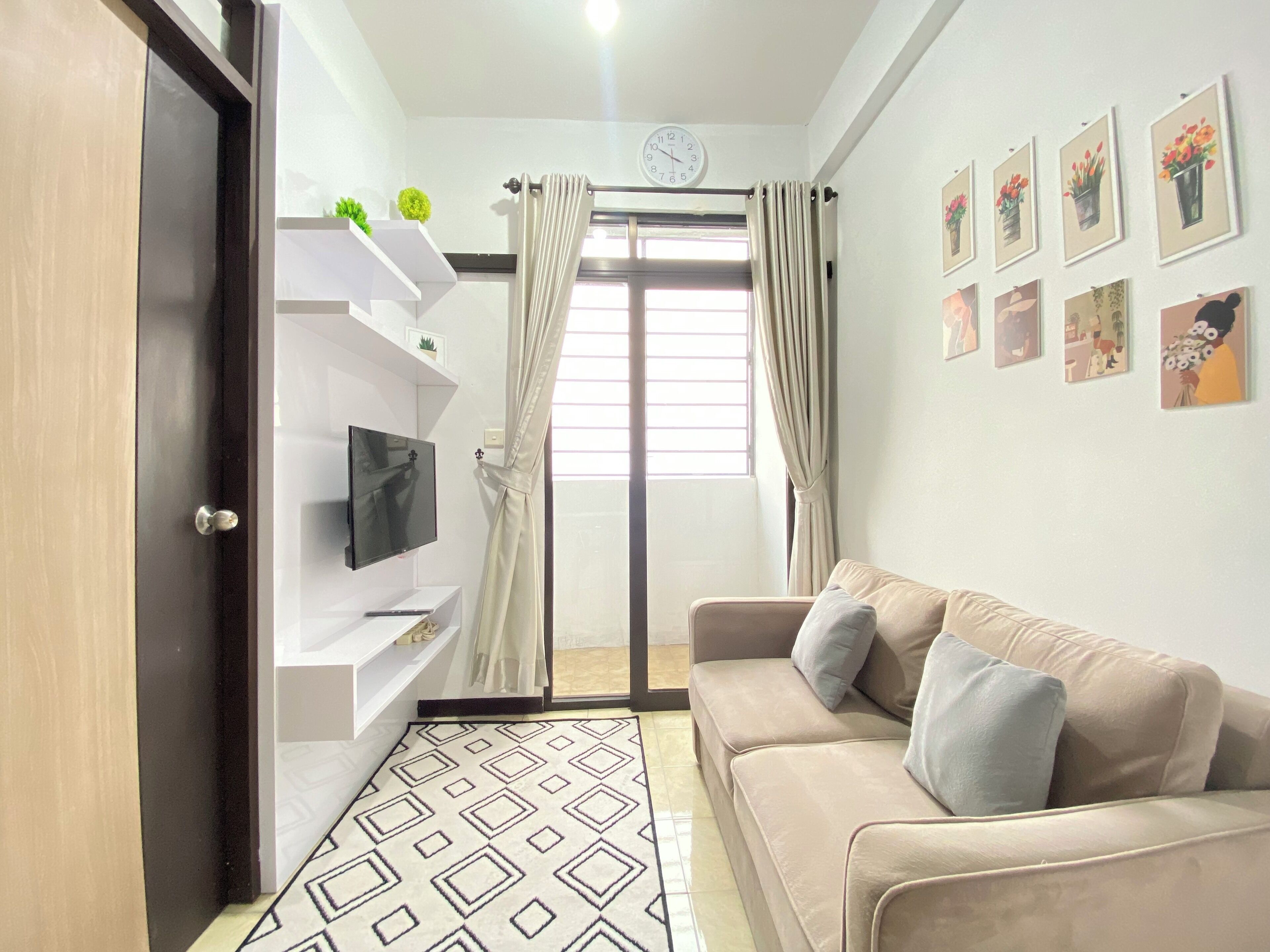 Un salon avec un canapé beige, un téléviseur à écran plat posé sur une étagère blanche, un tapis à motifs et une grande fenêtre avec des rideaux.