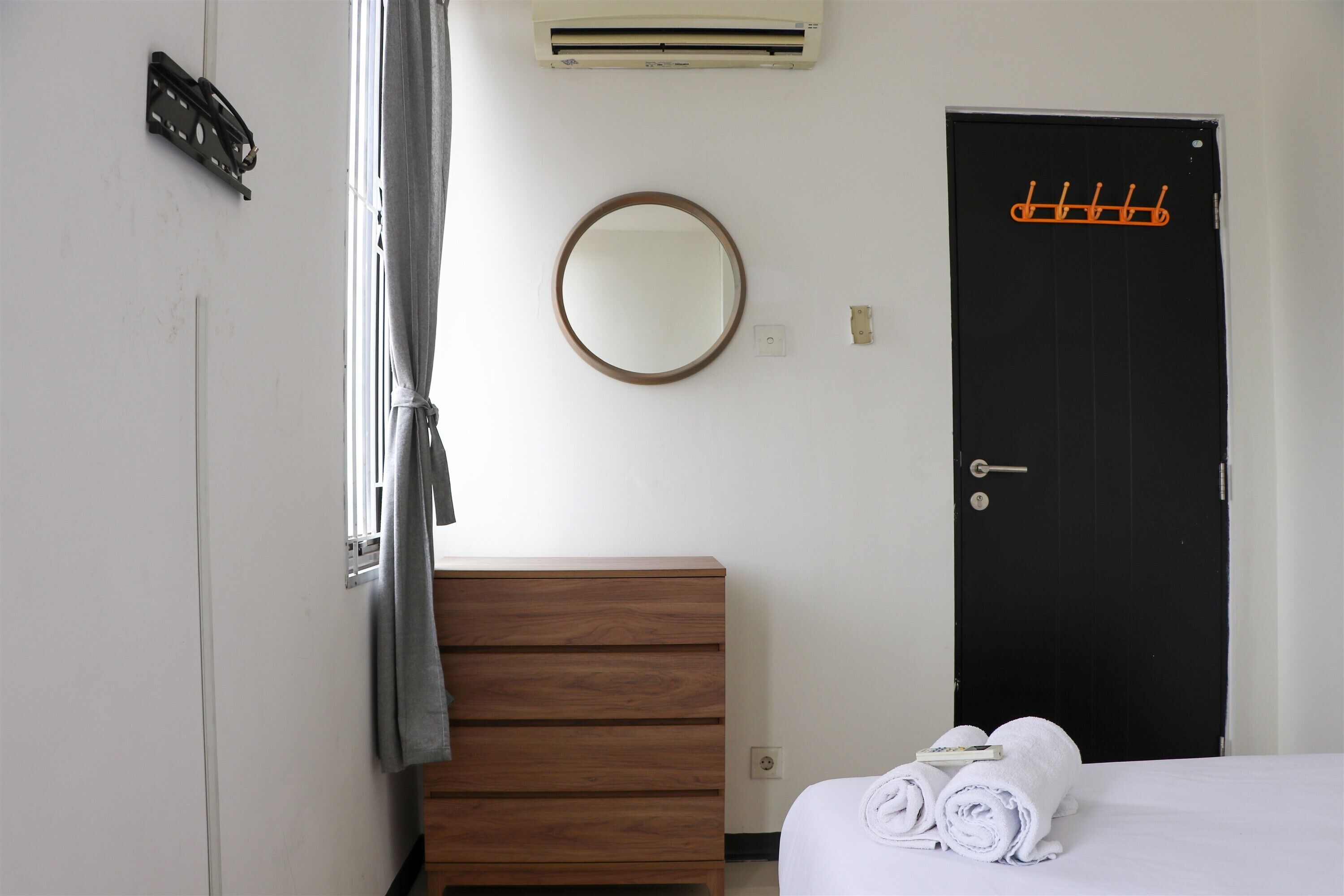 Ein Schlafzimmer mit einem hölzernen Schminktisch, einem runden Spiegel, einer schwarzen Tür mit Kleiderhaken und einem Fenster mit Vorhang.