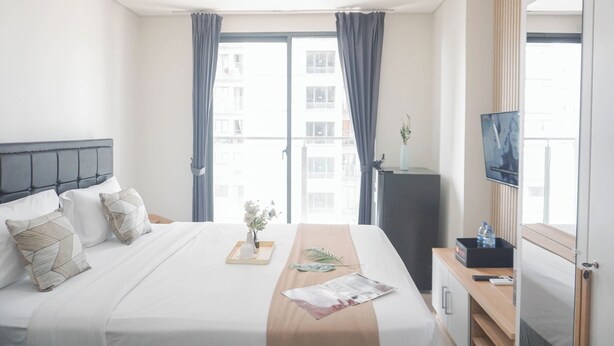 A hotel room with a bed, a bedside table with a tray, a plant, a magazine, a TV, and a window with curtains. - Full Furnished And Homey Studio At Daan Mogot City Apartment (Jakarta)