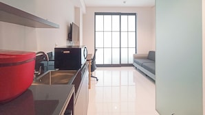 A modern living room with a large window, a flat-screen TV, a red storage bin, and a grey sofa. - Cozy And Enjoy Living Studio (No Kitchen) Apartment At Skales Residence Nusa Dua Bali (Nusa Dua)