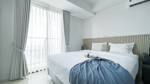 A modern hotel room with a large bed, white bedding, and a view of the city through sheer curtains. - Brand New And Comfortable Studio At Daan Mogot City Apartment (Jakarta)