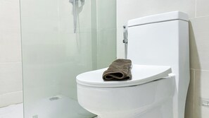 A white toilet with a towel on the seat, a glass shower enclosure, and a white tiled wall. - Brand New And Comfortable Studio At Daan Mogot City Apartment (Jakarta)