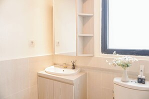 A bathroom with a white sink, a cabinet, a window, and a toilet with a vase of flowers. - Best Deal And Cozy 2Br Apartment At Meikarta (Cikarang)