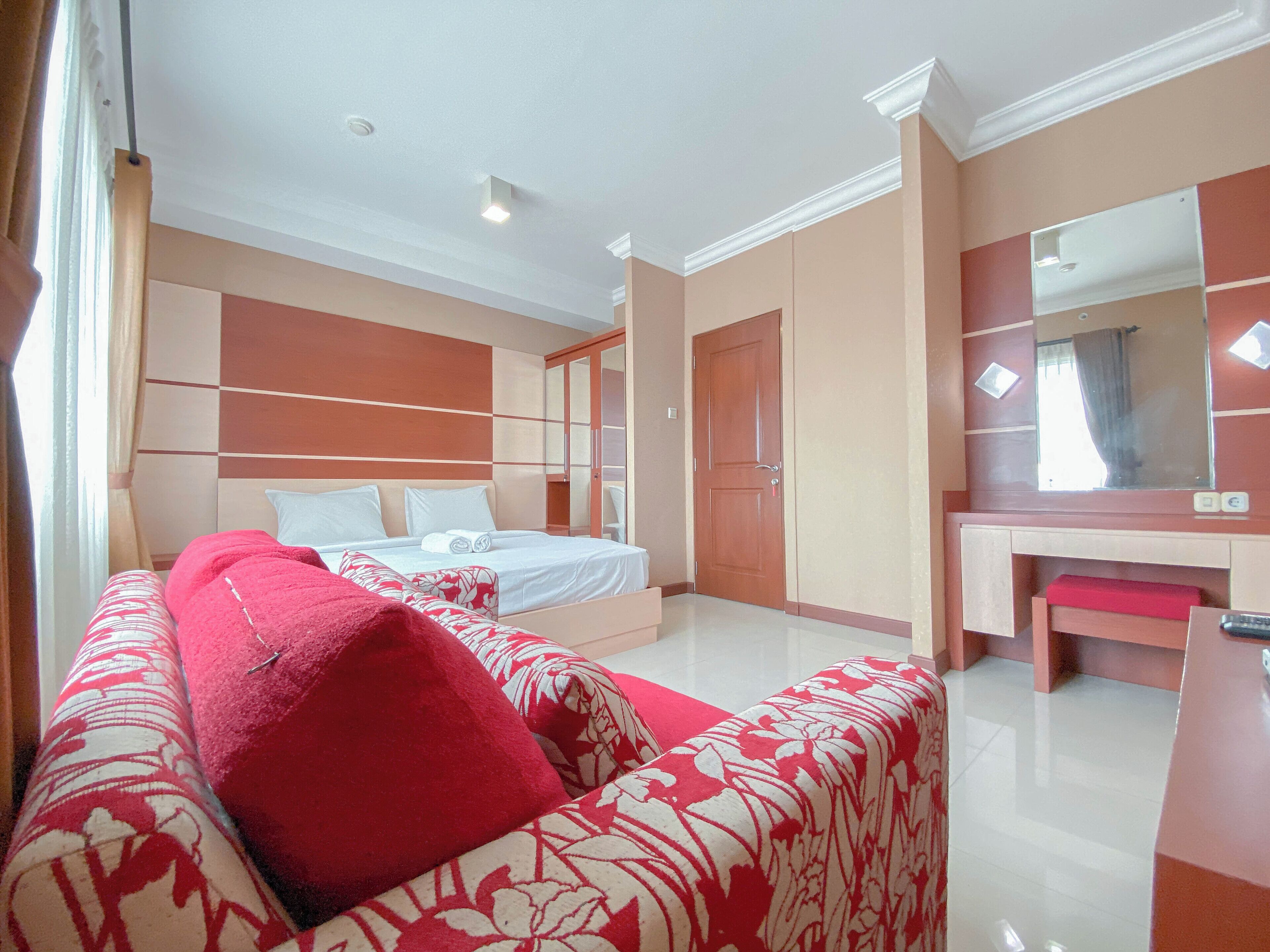 A bedroom with a bed, a red and white patterned sofa, a large mirror, and a wardrobe.