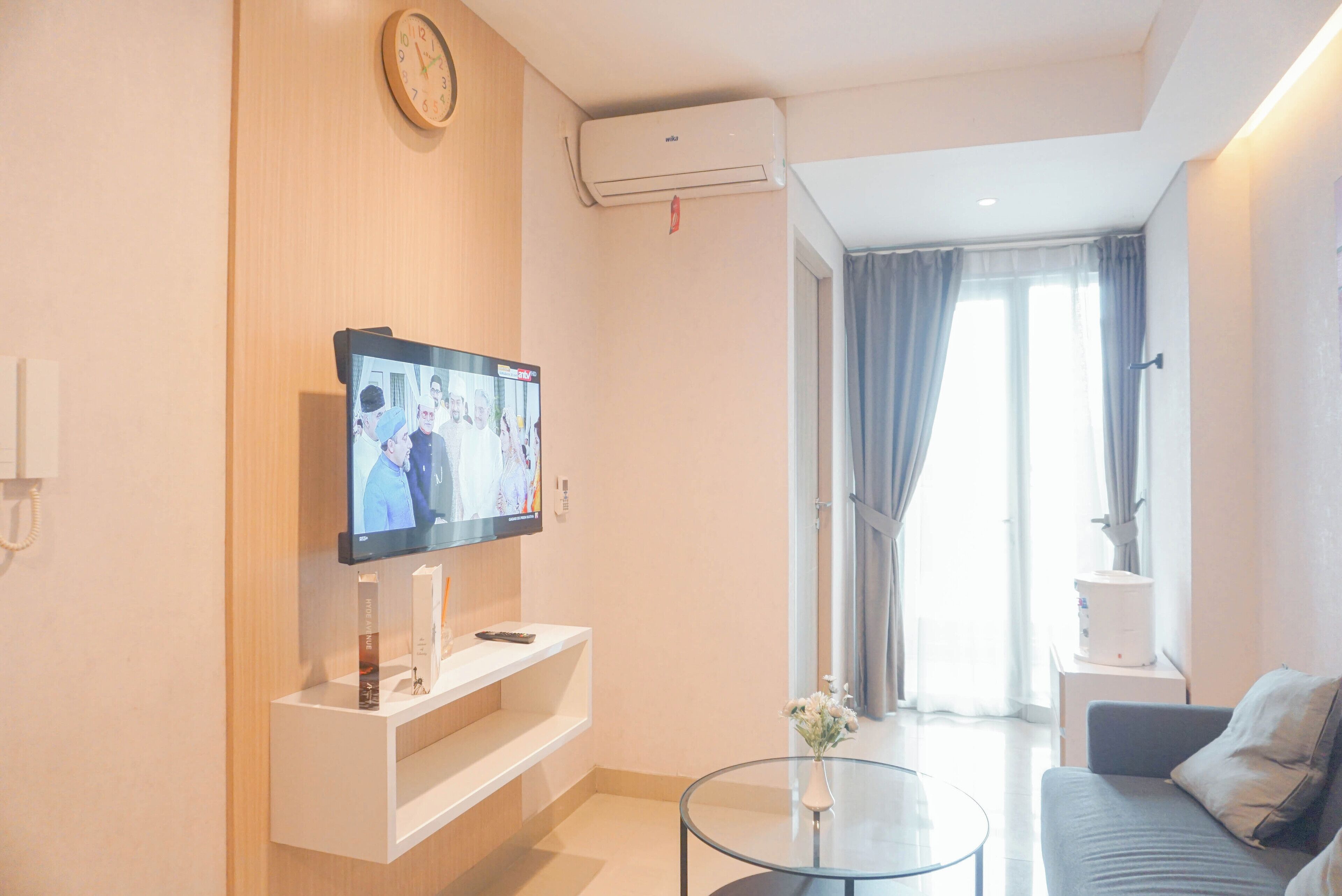 Un soggiorno moderno con una TV a schermo piatto montata su una parete in legno, un tavolino in vetro, un divano grigio e una finestra con tende.