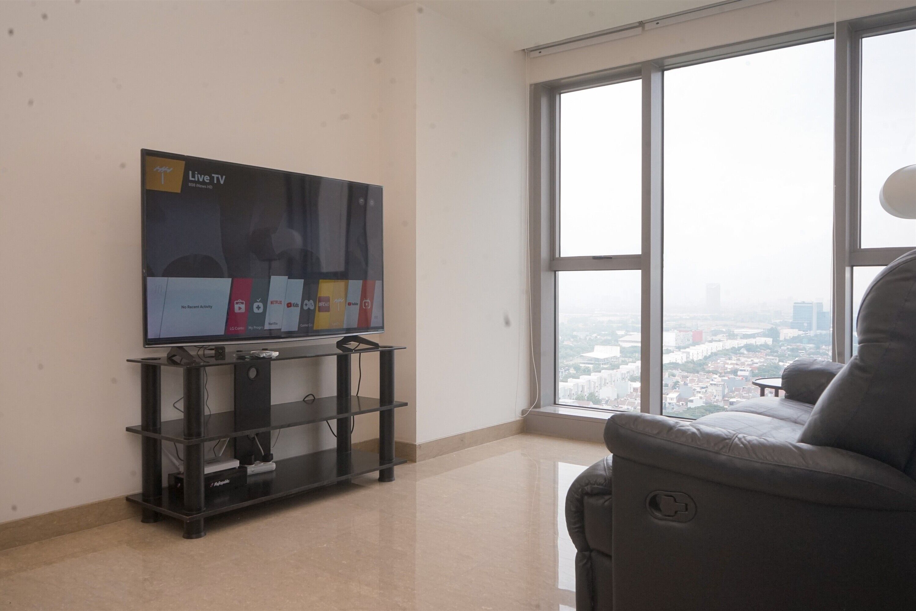 Ein Wohnzimmer mit einem großen Flachbildfernseher auf einem schwarzen Standfuß, einem grauen Sofa und einem großen Fenster mit Blick auf die Stadt.