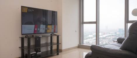 Ein Wohnzimmer mit einem großen Flachbildfernseher auf einem schwarzen Standfuß, einem grauen Sofa und einem großen Fenster mit Blick auf die Stadt.
