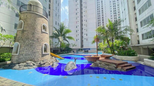 A swimming pool with a castle-like structure, palm trees, and a tall building in the background. - Best And Homey 2Br At Bassura City Apartment (Jakarta)