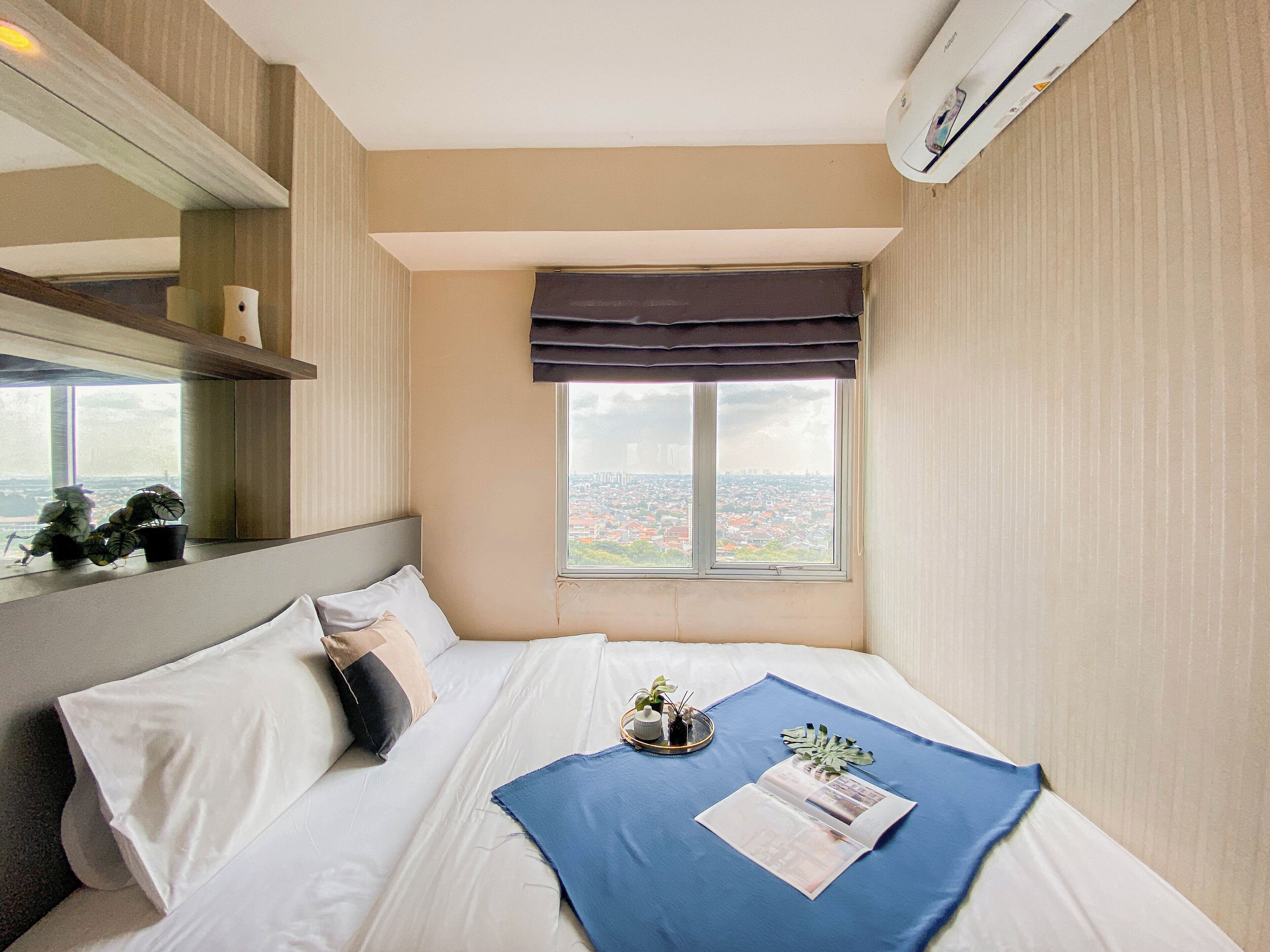 Una camera d'albergo con un letto, una finestra vista città, un comodino con una pianta e un condizionatore d'aria a parete.