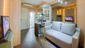 A modern living room with a white sofa, a TV, and a kitchen area with a tiled backsplash. - Best And Homey 2Br At Bassura City Apartment (Jakarta)
