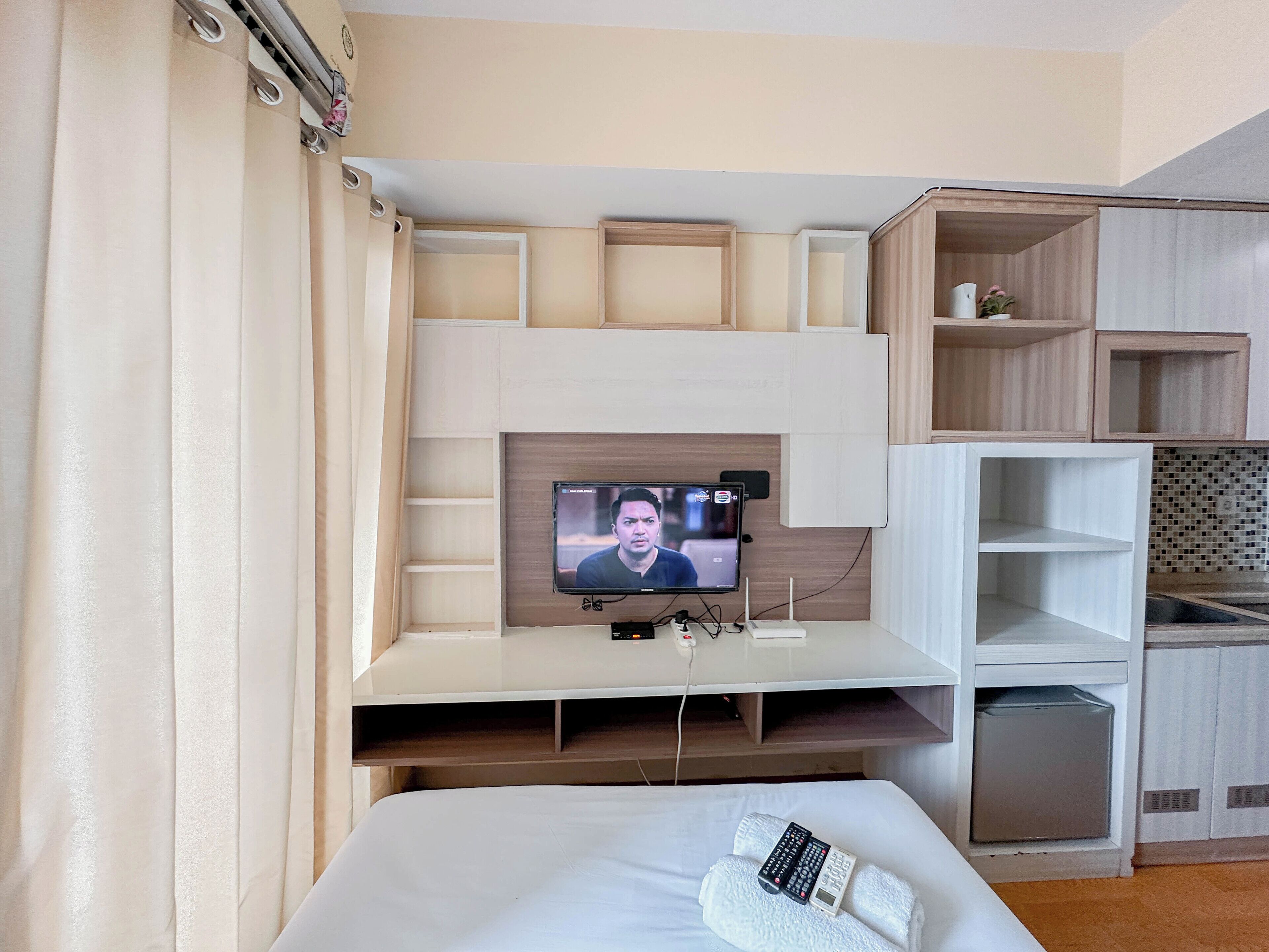 A hotel room with a bed, a TV mounted on a wooden wall, and a small shelf with a remote control.