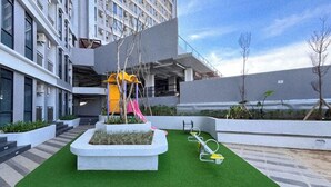 A playground with a slide and climbing structures, surrounded by planters and a green artificial turf area. - The Good Estate Studio At Delft Ciputra Makassar Apartment (Makassar)