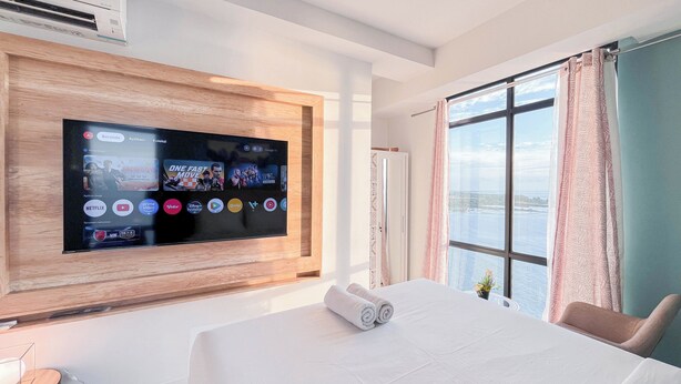 A hotel room with a large flat-screen TV mounted on a wooden panel, a bed with a white comforter, a window with curtains, and a view of the sea. - The Simply Look 1Br Apartment At Delft Ciputra Makassar (Makassar)