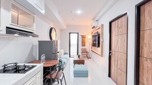 A modern kitchen with a stove, refrigerator, and dining area with chairs and a table. There is a hallway leading to a living area with a sofa and a TV mounted on the wall. - The Simply Look 1Br Apartment At Delft Ciputra Makassar (Makassar)