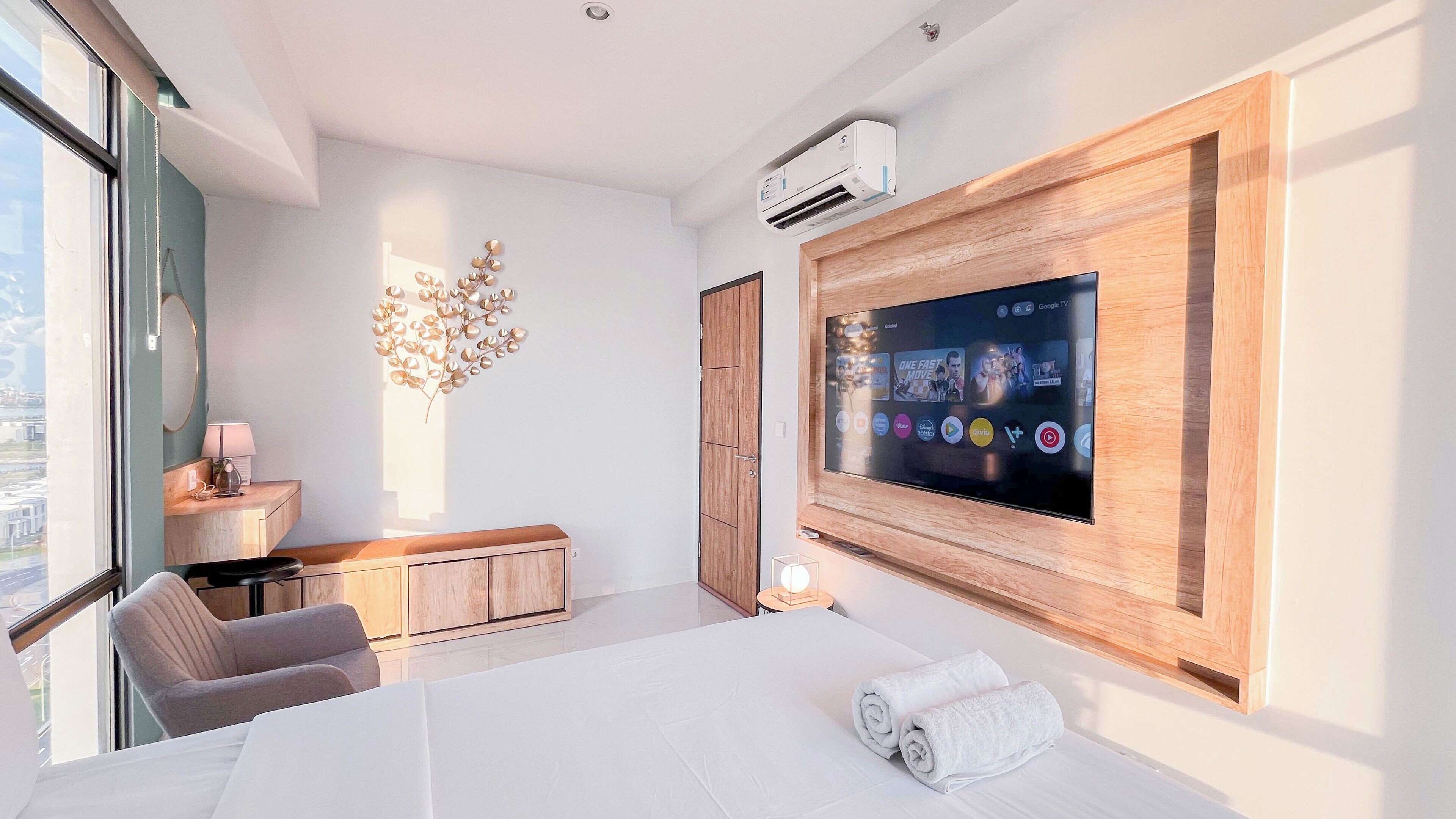 A modern hotel room with a large flat-screen TV mounted on the wall, a wooden cabinet, a desk with a lamp, a chair, and a bed with towels.