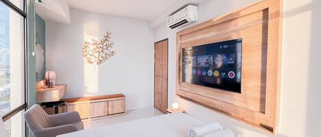 A modern hotel room with a large flat-screen TV mounted on the wall, a wooden cabinet, a desk with a lamp, a chair, and a bed with towels.