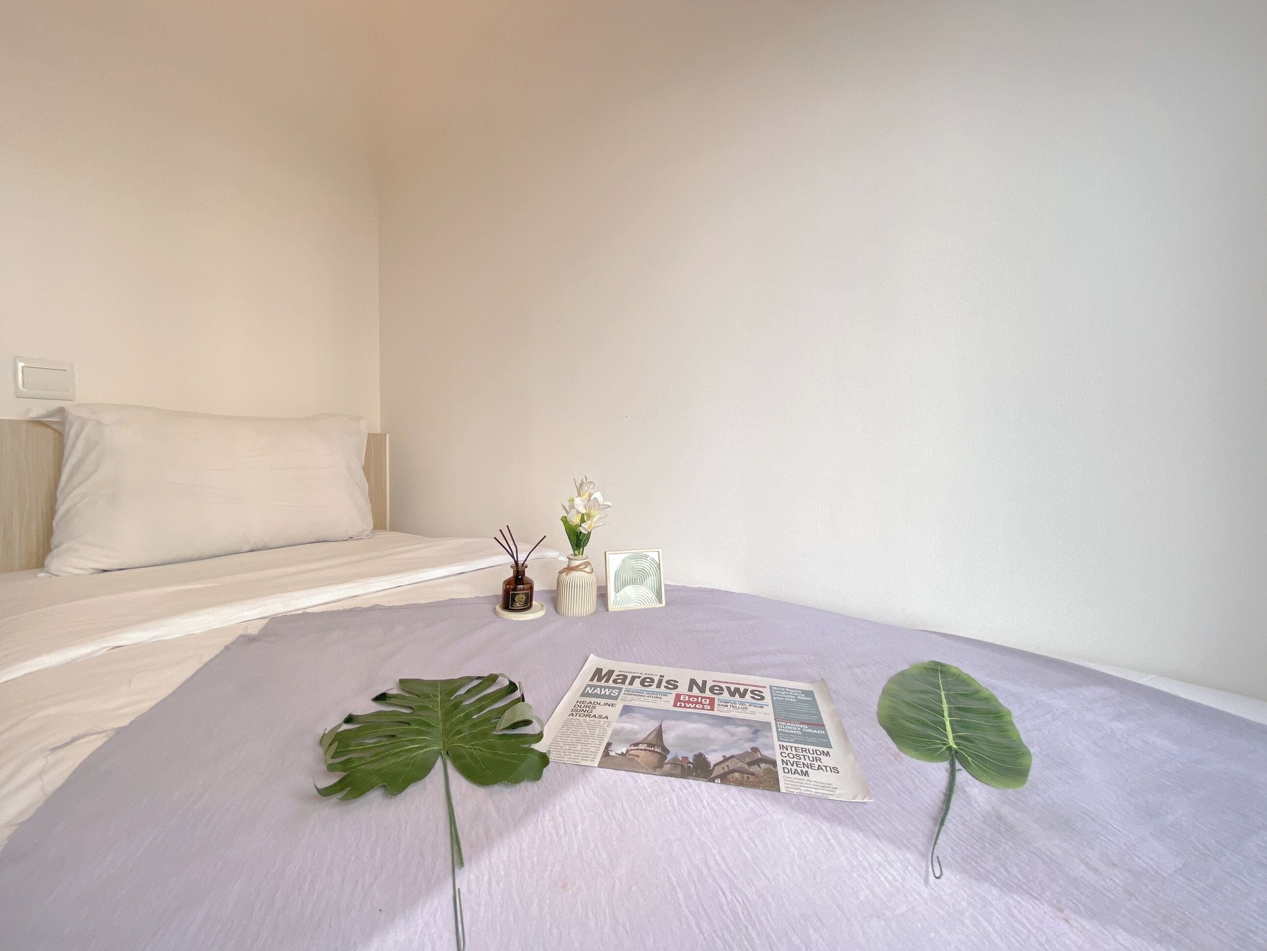 Ein ordentlich bezogenes Bett mit einem weißen Kopfkissen und einer weißen Daunendecke, ein kleiner Blumenstrauß in einer Vase, ein gerahmtes Bild und zwei große grüne Blätter auf dem Bett.