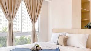 A neatly made bed with a blue throw blanket and a book, a wooden headboard, and a view of a building through sheer curtains. - Cozy Living 2Br Apartment Tokyo Riverside (Teluknaga)