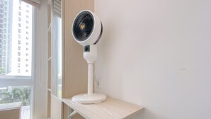A white standing fan on a wooden shelf in a room with a window. - Cozy Living 2Br Apartment Tokyo Riverside (Teluknaga)