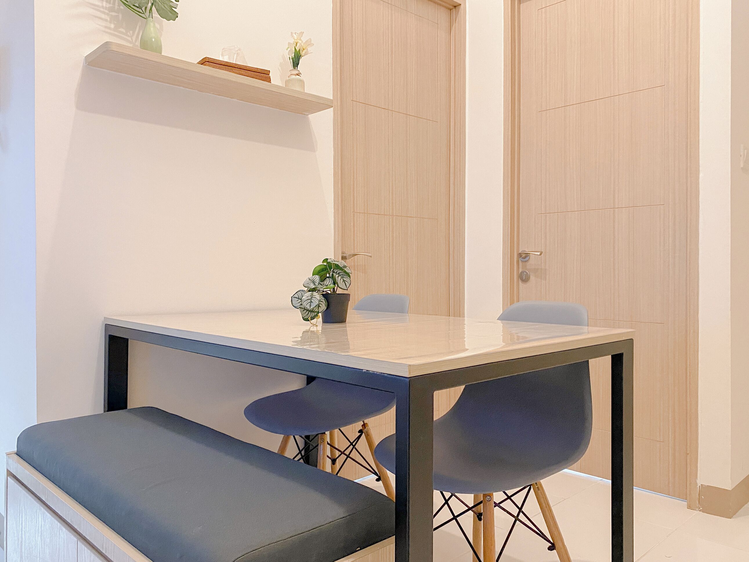 Ein moderner Essbereich mit einem rechteckigen Tisch, zwei blauen Stühlen und einer Bank. Über dem Tisch befindet sich ein Regal mit dekorativen Gegenständen.