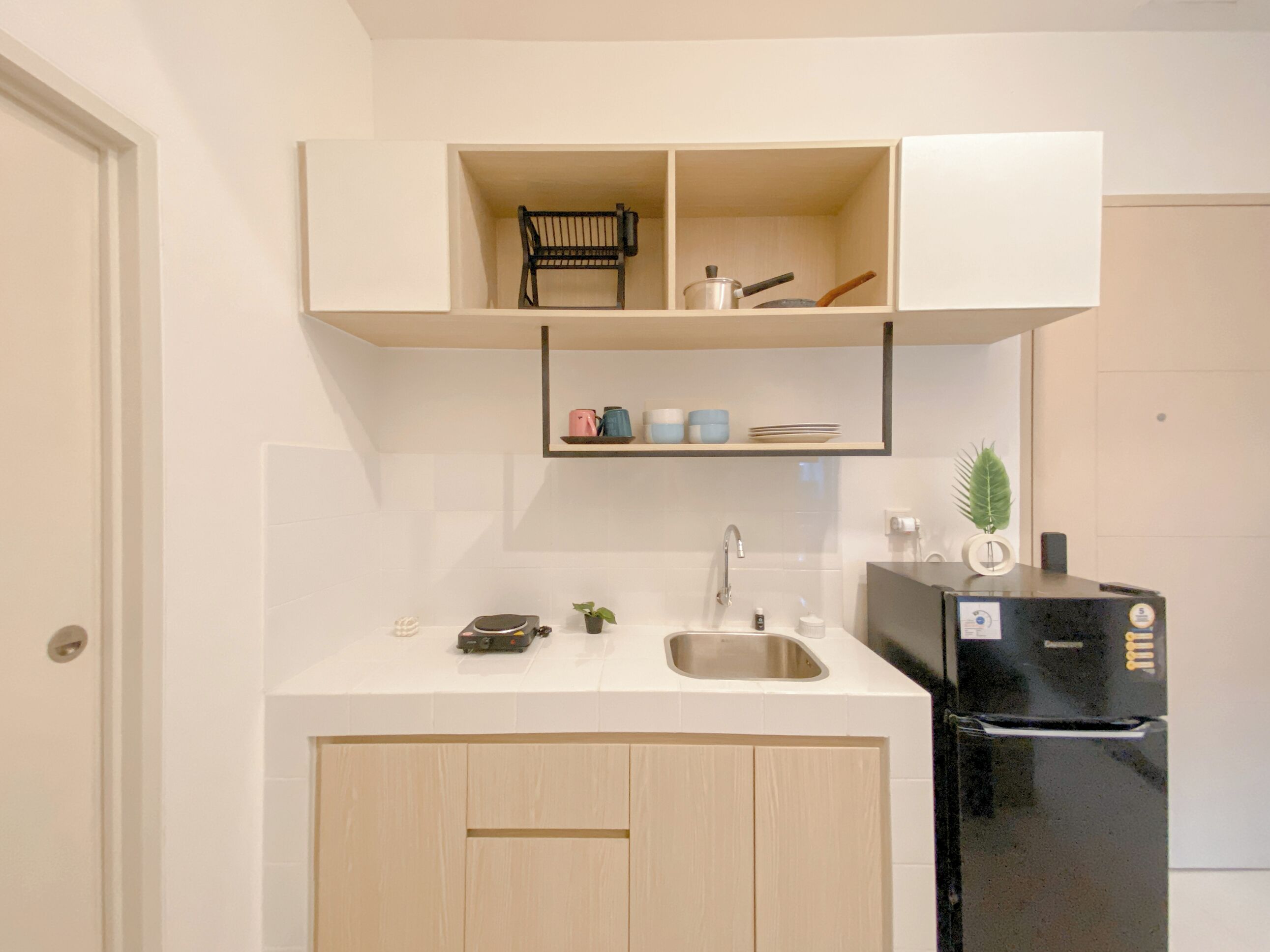 A compact kitchen with a built-in sink, a black refrigerator, and a shelf above the counter holding kitchenware and a plant.