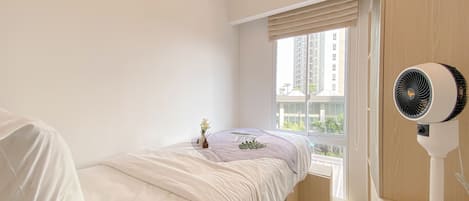 A bedroom with a bed, a window with blinds, a standing fan, and a small plant on the windowsill.