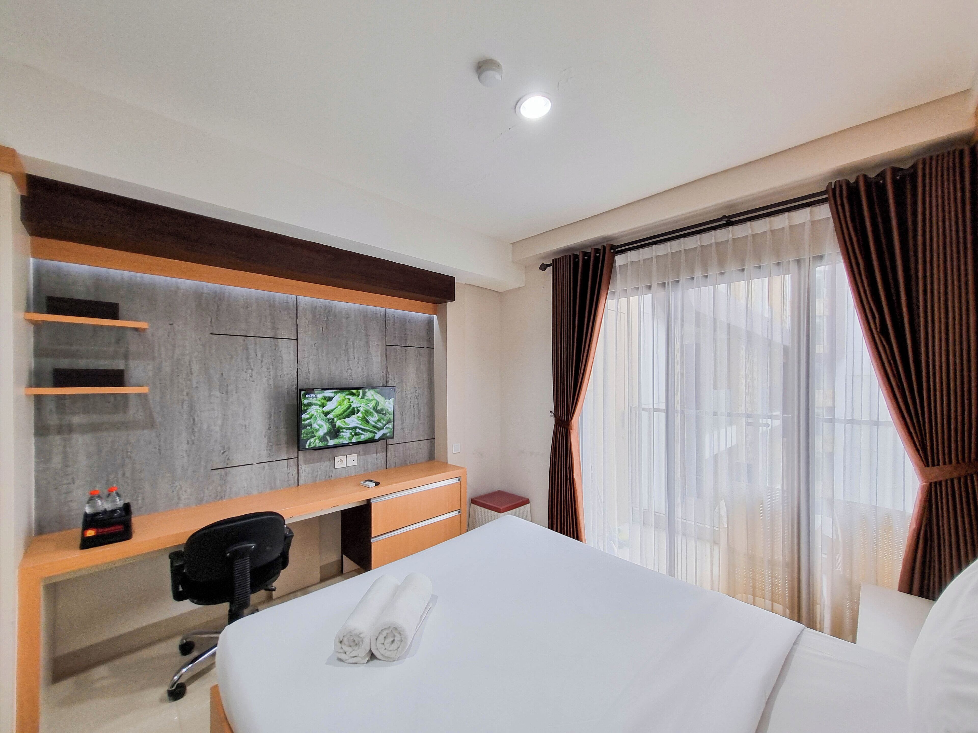 A hotel room with a bed, a desk with a computer, a TV mounted on the wall, and a window with curtains.