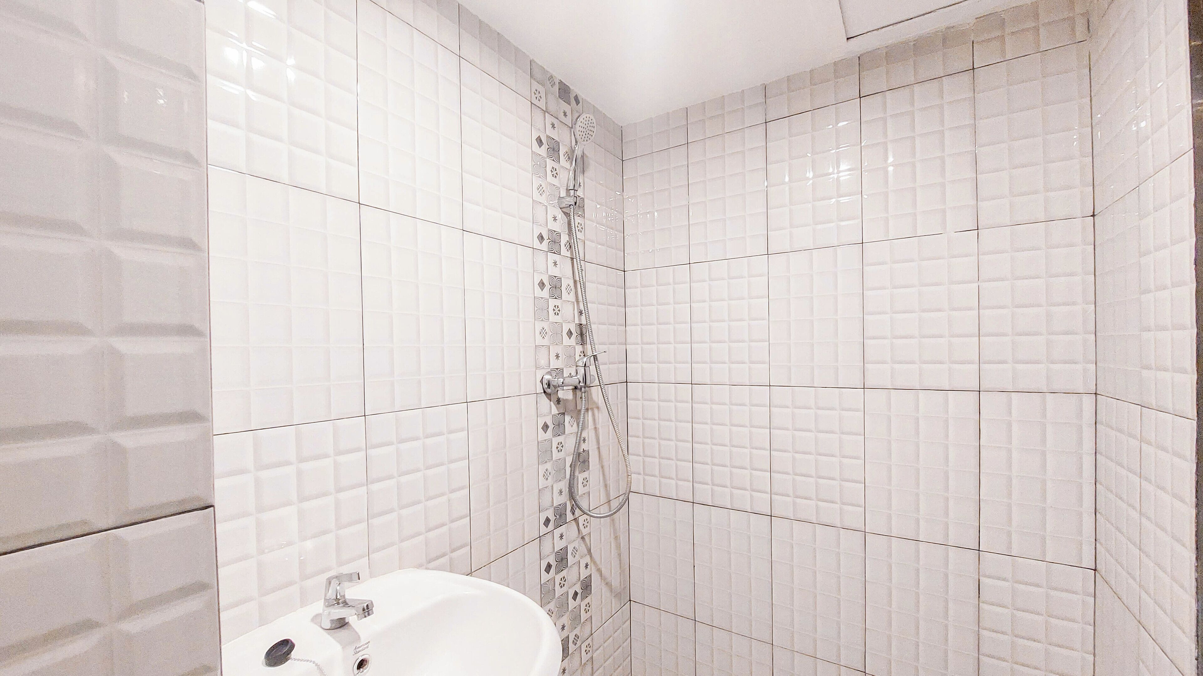 Un baño con paredes de azulejos blancos, una ducha con cabezal de ducha manual y un lavabo con grifo.
